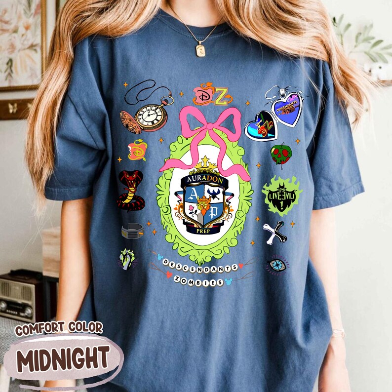 Może przedstawiać: Granatowa koszulka z kolorową grafiką przedstawiającą herb szkoły, r&oacute;żową kokardę i r&oacute;żne tematyczne zawieszki. Tekst "Descendants Zombies" jest wydrukowany na dole. Koszulka jest oznaczona jako "Comfort Color".