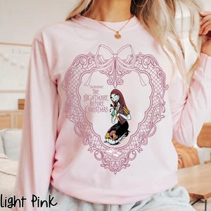 May include: Pale pink long-sleeved sweatshirt with a heart-shaped design and a bow. The design features the text "The Nightmare Before Christmas" and an illustration of Sally. The sweatshirt is a light pink colour.