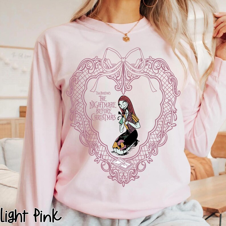 May include: Pale pink long-sleeved sweatshirt with a heart-shaped design and a bow. The design features the text "The Nightmare Before Christmas" and an illustration of Sally. The sweatshirt is a light pink colour.