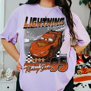 May include: A light purple t-shirt featuring a graphic of Lightning McQueen from the movie Cars. The design includes the text "Lightning McQueen", "Piston Cup Racing Series", and the number "95". The Pixar logo is also visible.