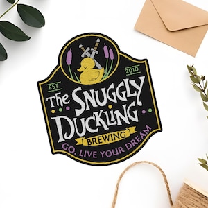 May include: Black and white logo with a yellow rubber ducky wearing a crown of swords. The text reads "The Snuggly Duckling Brewing Est. 2010 Go, Live Your Dream."