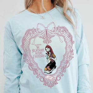 May include: Pale blue long-sleeved top with a pink heart-shaped design, a bow, and the text "Tim Burton's The Nightmare Before Christmas." The design features an illustration of Sally from the film.