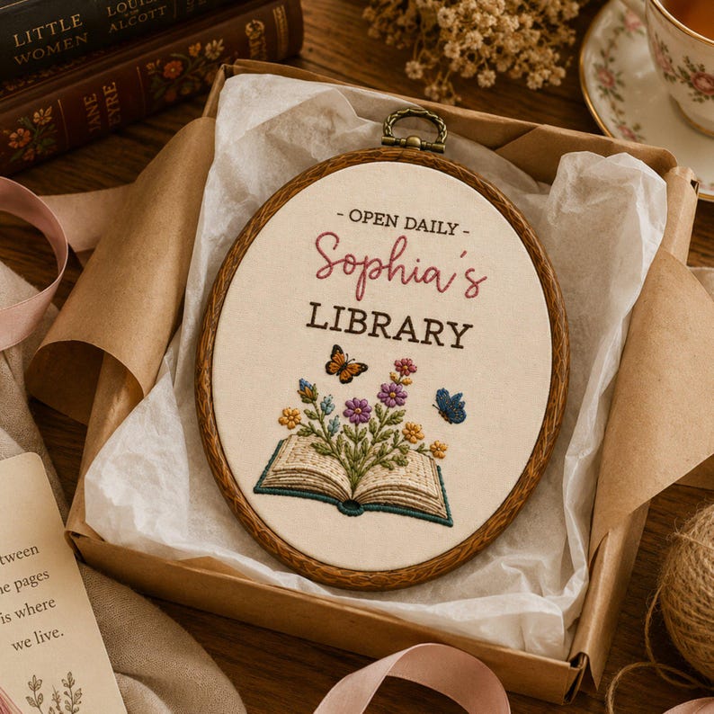 May include: An oval embroidery hoop containing cream fabric with the embroidered text OPEN DAILY Sophia's LIBRARY. Below the text is an open book with colorful flowers and green leaves emerging from the pages, accompanied by two small butterflies.
