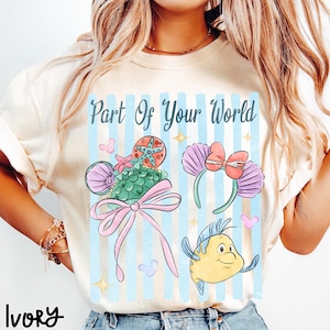 Puede incluir: Una camiseta color marfil con el texto Part Of Your World en letra cursiva azul oscuro. El gr&aacute;fico incluye rayas verticales azul claro, una diadema de orejas con patr&oacute;n de conchas verdes y lazo rosa, una diadema de conchas moradas y un pez amarillo.