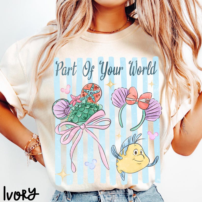 Puede incluir: Una camiseta color marfil con el texto Part Of Your World en letra cursiva azul oscuro. El gr&aacute;fico incluye rayas verticales azul claro, una diadema de orejas con patr&oacute;n de conchas verdes y lazo rosa, una diadema de conchas moradas y un pez amarillo.