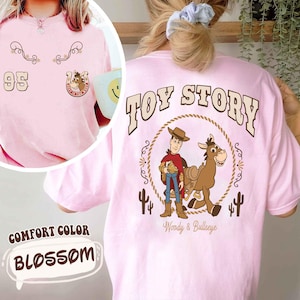 May include: A light pink t-shirt with the text "TOY STORY" on the back, featuring Woody and Bullseye. The front has the number "95" and a horseshoe graphic. The shirt is labeled "Comfort Color Blossom".