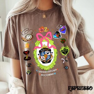 Może przedstawiać: Brązowa koszulka z kolorowym wzorem graficznym przedstawiającym herb szkoły, r&oacute;żową kokardę i r&oacute;żne ilustracje tematyczne. Widoczny jest tekst "AURADON PREP" wraz ze słowem "DESCENDANTS ZOMBIES". Słowo "ESPRESSO" jest wydrukowane na dole.