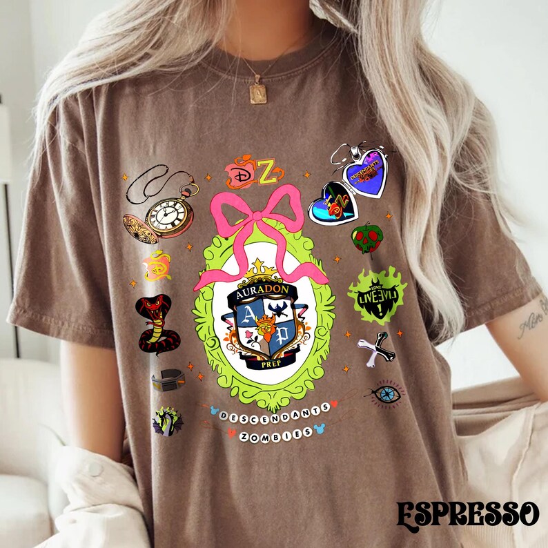 Może przedstawiać: Brązowa koszulka z kolorowym wzorem graficznym przedstawiającym herb szkoły, r&oacute;żową kokardę i r&oacute;żne ilustracje tematyczne. Widoczny jest tekst "AURADON PREP" wraz ze słowem "DESCENDANTS ZOMBIES". Słowo "ESPRESSO" jest wydrukowane na dole.