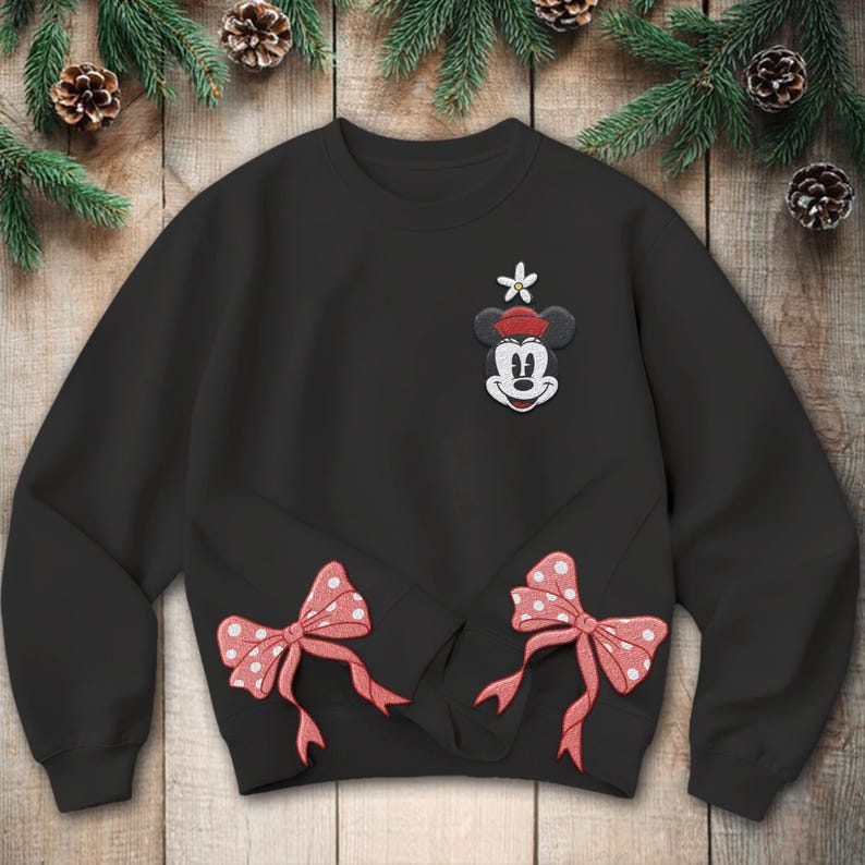 Może przedstawiać: Czarna bluza z naszywką Minnie Mouse z czerwoną kokardą i białymi kropkami. Dwa duże czerwone kokardy z białymi kropkami na dole. Projekt jest świąteczny i odpowiedni na sezon świąteczny.
