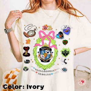 Może przedstawiać: Kremowa koszulka z kolorowym nadrukiem graficznym z napisem "Descendants Zombies". Projekt zawiera r&oacute;żową kokardę, zegar, węża i inne elementy tematyczne. Koszulka ma luźny kr&oacute;j.