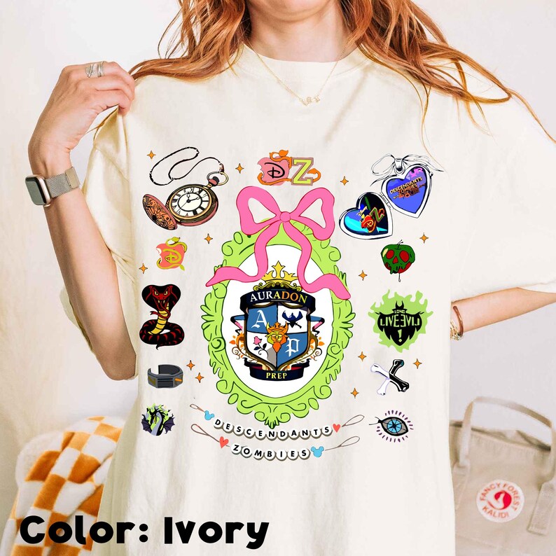 Może przedstawiać: Kremowa koszulka z kolorowym nadrukiem graficznym z napisem "Descendants Zombies". Projekt zawiera r&oacute;żową kokardę, zegar, węża i inne elementy tematyczne. Koszulka ma luźny kr&oacute;j.