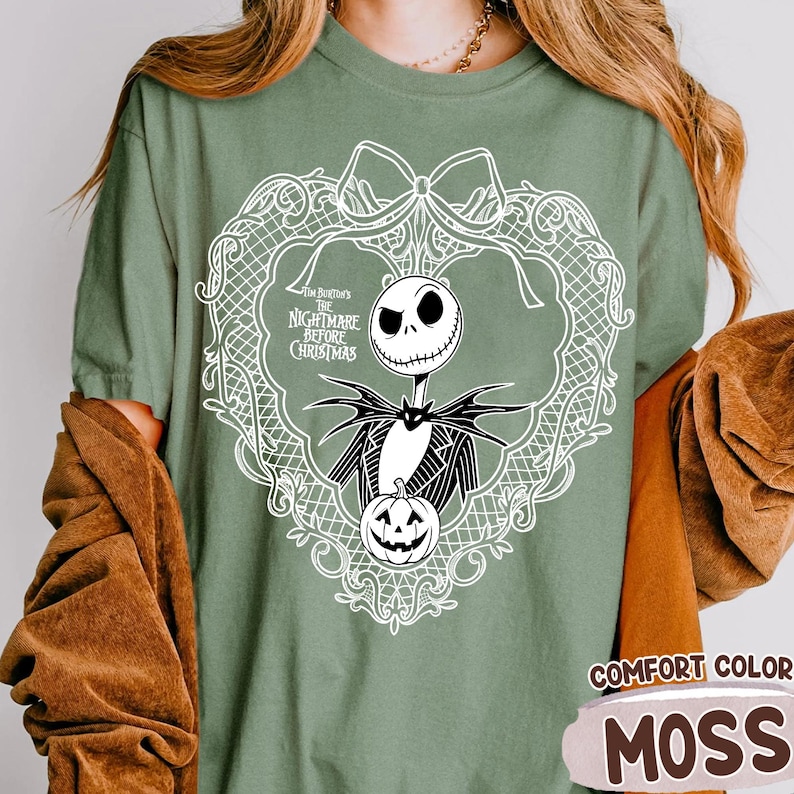 May include: Moss green t-shirt with a white graphic of Jack Skellington from "The Nightmare Before Christmas" in a decorative frame. The shirt also has the words "Comfort Color" and "Moss" printed on it.