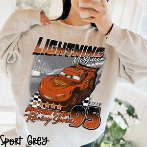 May include: Sport grey sweatshirt featuring a graphic of Lightning McQueen from the movie Cars. The design includes the text "Lightning McQueen", "Piston Cup Racing Series", and the number "95".