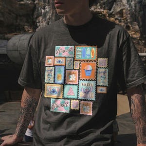 May include: Dark gray t-shirt featuring a collage of colorful postage stamp designs with Hawaiian themes. The stamps include images of Stitch, pineapples, surfboards, and the word "Hawaii". The shirt has a relaxed fit.