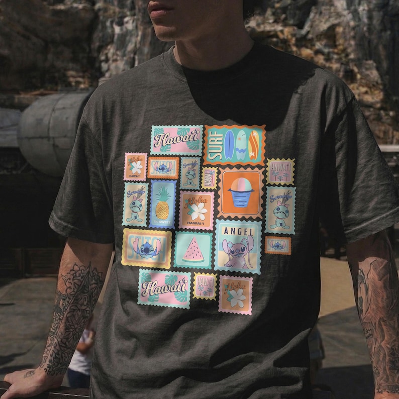 May include: Dark gray t-shirt featuring a collage of colorful postage stamp designs with Hawaiian themes. The stamps include images of Stitch, pineapples, surfboards, and the word "Hawaii". The shirt has a relaxed fit.