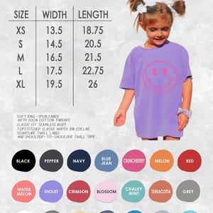 May include: A lilac youth t-shirt with a large pink smiley face graphic. The image includes a size chart with measurements in inches, and a color palette with options like black, navy, and red. The shirt is made of soft ring-spun fabric.