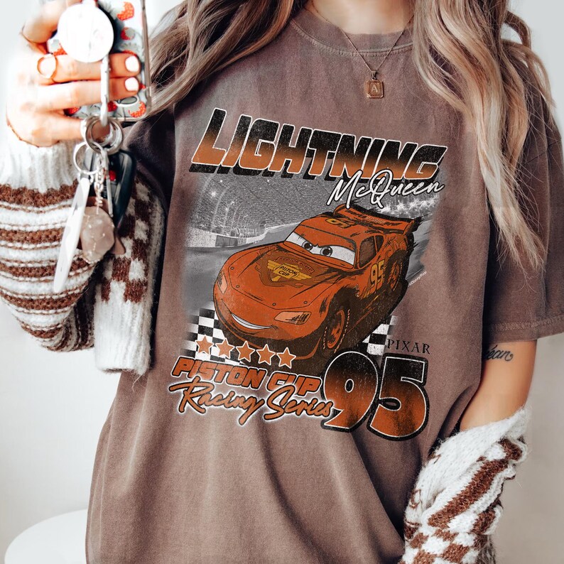 May include: Brown t-shirt featuring a graphic of Lightning McQueen from the movie Cars. The design includes the text "LIGHTNING McQueen", "PIXAR", "95", and "Piston Cup Racing Series". The shirt has a vintage look.