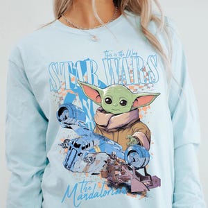 May include: Light blue long-sleeve shirt featuring a graphic of Grogu from Star Wars, with the text "This is the Way" and "Star Wars" in a stylized font. The Mandalorian and a blue ship are also depicted.