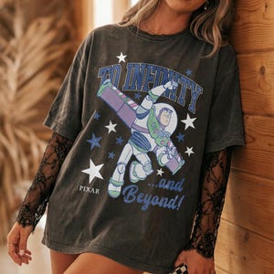 Puede incluir: Camiseta gris oscuro con Buzz Lightyear de Toy Story de Pixar. El gr&aacute;fico incluye la frase "To Infinity...and Beyond!" con acentos de estrellas. La camiseta tiene mangas cortas y se usa con una manga de encaje negro debajo.
