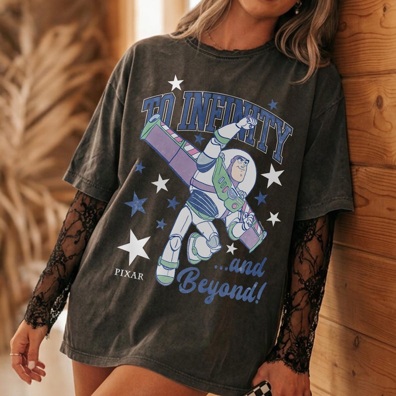 Puede incluir: Camiseta gris oscuro con Buzz Lightyear de Toy Story de Pixar. El gr&aacute;fico incluye la frase "To Infinity...and Beyond!" con acentos de estrellas. La camiseta tiene mangas cortas y se usa con una manga de encaje negro debajo.