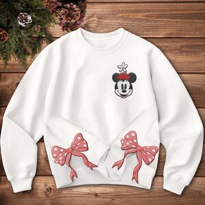 Może przedstawiać: Biała bluza z naszywką Myszki Miki w czerwonym kapeluszu i kwiatkiem. Dwie czerwone kokardy w kropki są wyhaftowane na dolnej przedniej części. Bluza leży na drewnianej powierzchni.