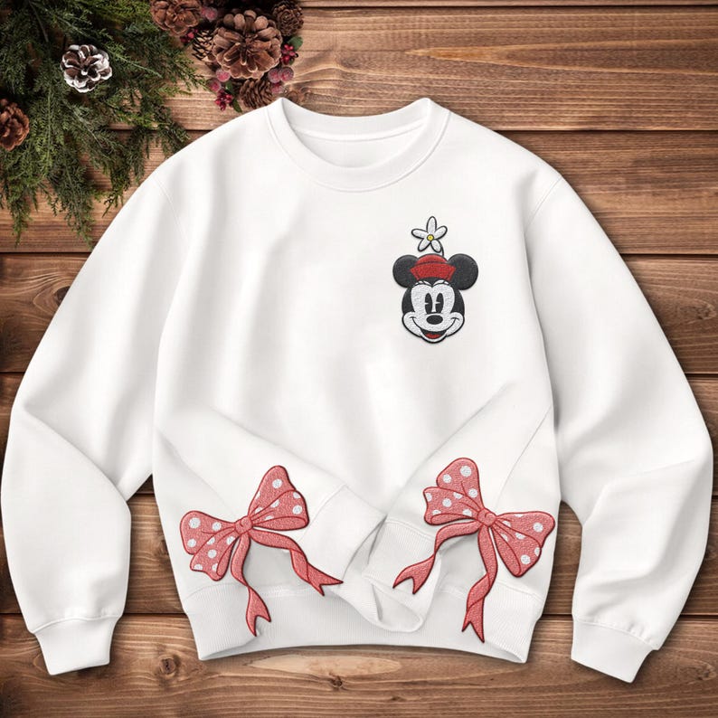 Może przedstawiać: Biała bluza z naszywką Myszki Miki w czerwonym kapeluszu i kwiatkiem. Dwie czerwone kokardy w kropki są wyhaftowane na dolnej przedniej części. Bluza leży na drewnianej powierzchni.