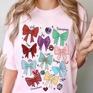 May include: A light pink t-shirt featuring a colorful graphic of bows with names like "Mal," "Jay," and "Descendants." The bows are in various colors, including red, purple, green, and pink, with decorative elements.