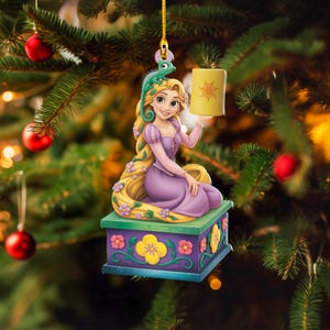 May include: A Christmas tree ornament featuring a cartoon character with long blonde hair, wearing a purple dress, and holding a yellow lantern. A green chameleon sits on her head. The ornament is on a decorative box.