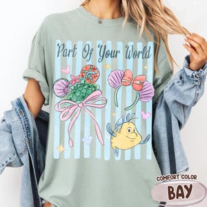Puede incluir: Una camiseta verde salvia con el texto Part Of Your World en letra azul oscuro. El gr&aacute;fico incluye una diadema de sirena verde con un lazo rosa, una diadema de conchas moradas con un lazo rojo y una ilustraci&oacute;n de un pez amarillo.