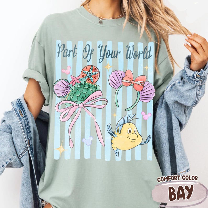 Puede incluir: Una camiseta verde salvia con el texto Part Of Your World en letra azul oscuro. El gr&aacute;fico incluye una diadema de sirena verde con un lazo rosa, una diadema de conchas moradas con un lazo rojo y una ilustraci&oacute;n de un pez amarillo.