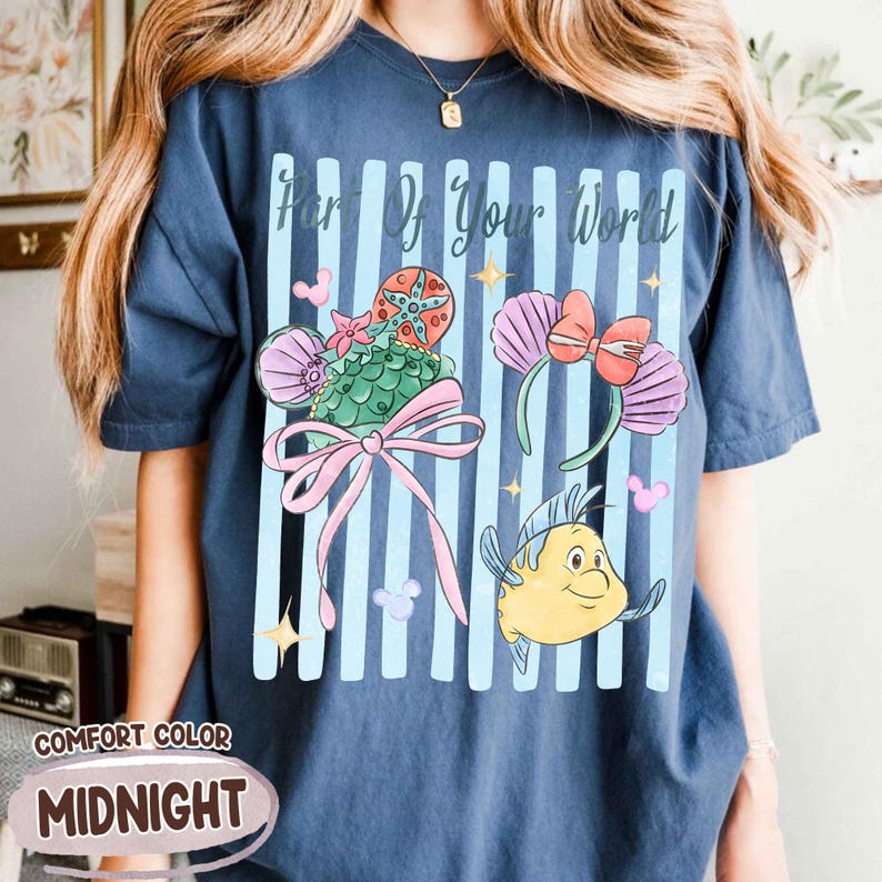 Puede incluir: Una camiseta azul marino con el texto Part Of Your World sobre un gr&aacute;fico de dos pares de orejas de rat&oacute;n, una con una concha verde y lazo rosa, la otra con conchas moradas y lazo rojo, junto a un personaje de pez amarillo y estrellas.