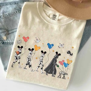 May include: A beige t-shirt with a graphic print of Star Wars characters wearing Mickey Mouse ears and holding balloons. The characters include Darth Vader, Stormtroopers, and a droid. The text "COMFORT COLORS" is visible on the shirt.