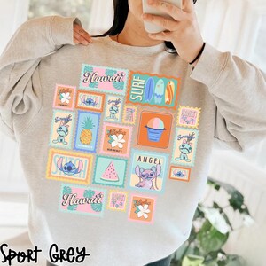 May include: Sport grey sweatshirt featuring a collage of colorful postage stamp designs with Hawaiian themes. The stamps include images of Stitch, pineapples, flowers, surfboards, and the words "Hawaii", "Angel", and "Surf".