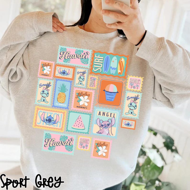 May include: Sport grey sweatshirt featuring a collage of colorful postage stamp designs with Hawaiian themes. The stamps include images of Stitch, pineapples, flowers, surfboards, and the words "Hawaii", "Angel", and "Surf".