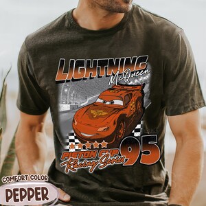 May include: Dark grey t-shirt featuring a graphic of Lightning McQueen from the movie Cars. The design includes the text "LIGHTNING McQueen", "Piston Cup Racing Series", and the number "95". The shirt is labeled "Comfort Color Pepper".