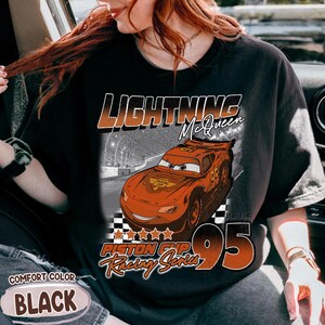 May include: Black t-shirt featuring Lightning McQueen from the movie Cars. The design includes the text "LIGHTNING McQueen", "Piston Cup Racing Series", and the number "95". The shirt is labeled "Comfort Color BLACK".