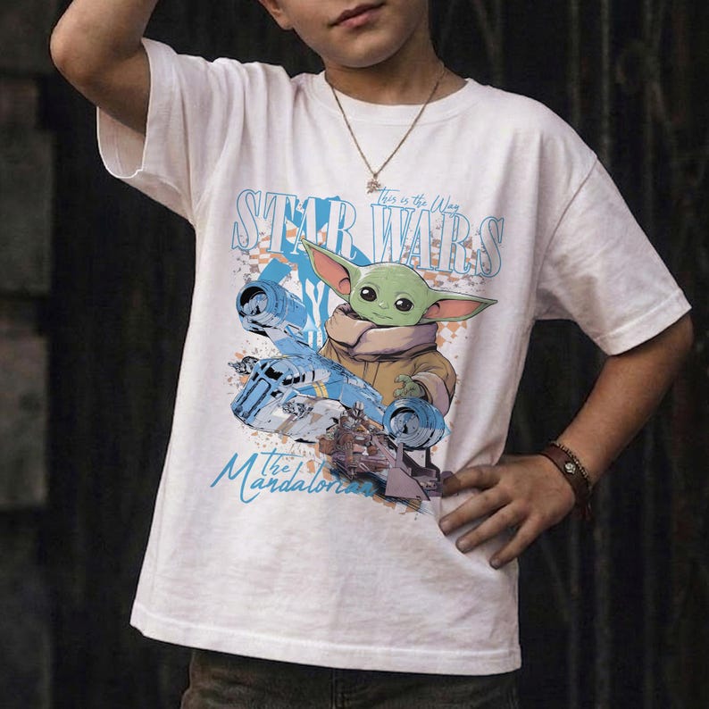 May include: White t-shirt featuring a graphic of Grogu from The Mandalorian series. The design includes the text "Star Wars" and "This is the Way" in blue, along with an image of Grogu and a blue spaceship. A brown bracelet is visible.