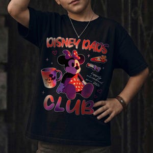 Może przedstawiać: Czarny t-shirt z grafiką Minnie Mouse i napisem "DISNEY DADS CLUB". Projekt zawiera kubek, spinkę do włos&oacute;w i frazę "happily outnumbered since 2020".