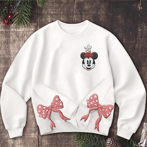 Może przedstawiać: Biała bluza z haftowanym wzorem Myszki Miki na g&oacute;rnej lewej piersi. Wz&oacute;r zawiera czerwony kapelusz i kwiat. Dwa czerwone łuki w kropki znajdują się na dole. Świąteczny element odzieży.