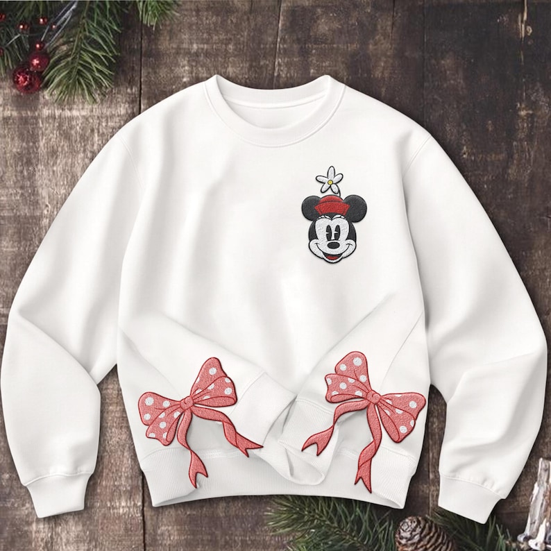 Może przedstawiać: Biała bluza z haftowanym wzorem Myszki Miki na g&oacute;rnej lewej piersi. Wz&oacute;r zawiera czerwony kapelusz i kwiat. Dwa czerwone łuki w kropki znajdują się na dole. Świąteczny element odzieży.