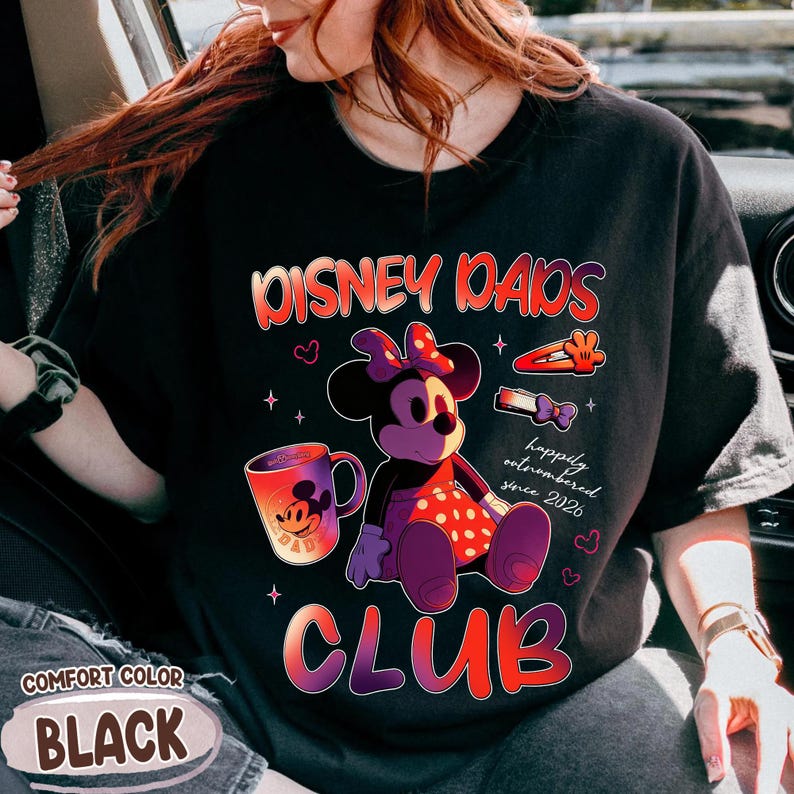 Może przedstawiać: Czarna koszulka z napisem "DISNEY DADS CLUB" w kolorze czerwonym i pomarańczowym. Koszulka przedstawia grafikę Minnie Mouse, kubek do kawy i frazę "happily outnumbered since 2026". Koszulka jest oznaczona jako "COMFORT COLOR BLACK".