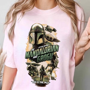 Puede incluir: Camiseta rosa claro con un dise&ntilde;o gr&aacute;fico de la serie Star Wars, "The Mandalorian y Grogu." El dise&ntilde;o incluye el casco del Mandaloriano, Grogu y la frase "This is the Way."