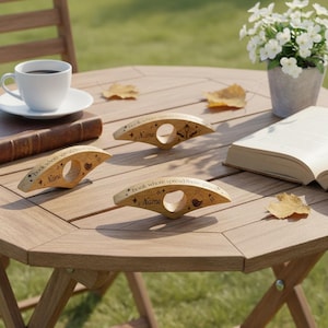 May include: Three wooden book holders with a circular finger hole and engraved text and designs are displayed on a wooden table. A cup of coffee, a book, and a small vase of white flowers are also on the table. The book holders have the text "book whore spread those pages" and the name "Name".