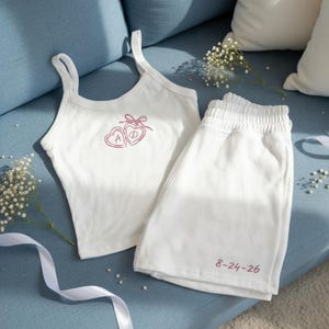 May include: White tank top and shorts set. The tank top features a pink heart design with the letters "A" and "D". The shorts have the date "8-24-26" printed in pink. The set is made of a soft material and is displayed on a blue couch.