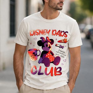 Może przedstawiać: Kremowa koszulka z grafiką Minnie Mouse i napisem "DISNEY DADS CLUB". Projekt zawiera kubek, spinkę do włos&oacute;w i frazę "happily outnumbered since 2026."