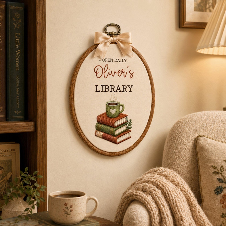 May include: An oval embroidery hoop wall hanging featuring a cream-colored fabric center with the text "OPEN DAILY Oliver's LIBRARY" in brown and black. Below the text is an illustration of three stacked books topped with a green coffee mug.