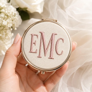 May include: A round compact mirror with a gold-toned metal frame and a cream-colored fabric cover. The center of the cover features the embroidered monogram letters EMC in a dusty rose thread. The mirror has a small hinge at the top and a clasp.