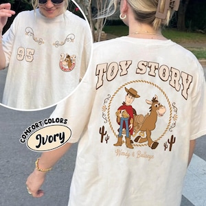 May include: Ivory-colored t-shirt featuring a "Toy Story" design on the back with Woody and Bullseye. The front has the number "95" and decorative elements. The shirt is labeled "Comfort Colors" and "Ivory".