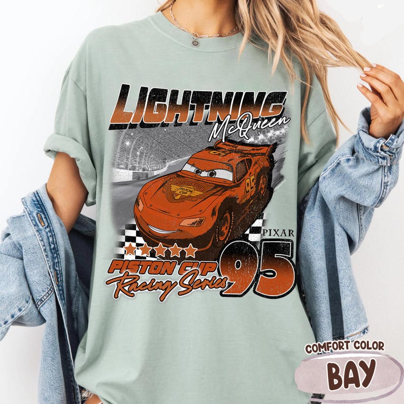 May include: A light green Comfort Color t-shirt featuring a graphic of Lightning McQueen from the Pixar Cars movie. The design includes the text "LIGHTNING McQueen", "PISTON CUP Racing Series", and the number "95". The shirt is labeled "COMFORT COLOR BAY".