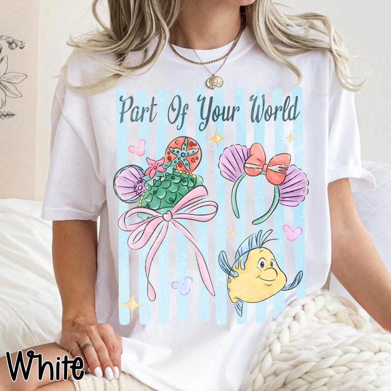 Puede incluir: Una camiseta blanca con el texto Part Of Your World en letra azul. El gr&aacute;fico incluye una concha verde con patr&oacute;n de escamas y un lazo rosa, una diadema de conchas moradas con un lazo rojo y un pez amarillo sobre rayas de color azul claro.